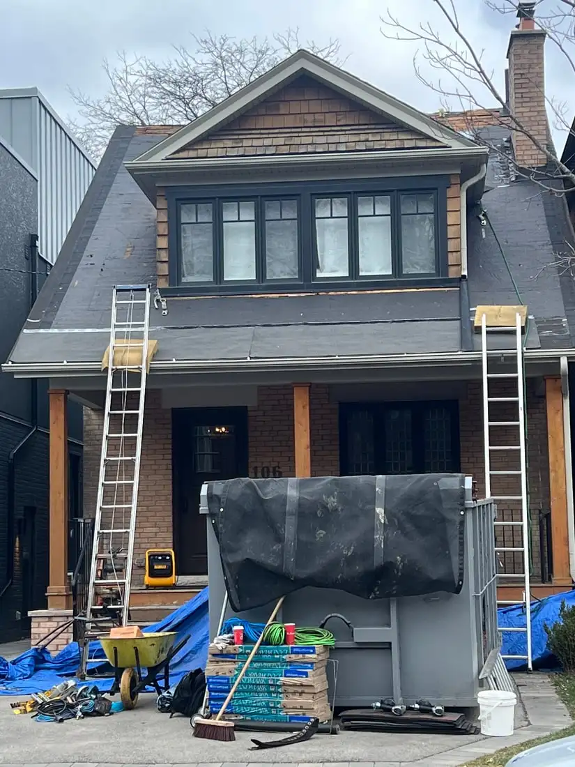 A brick and cedar-shingle craftsman two-storey stripped to black underlayment, two ladders up and a tarped rental bin in the driveway.