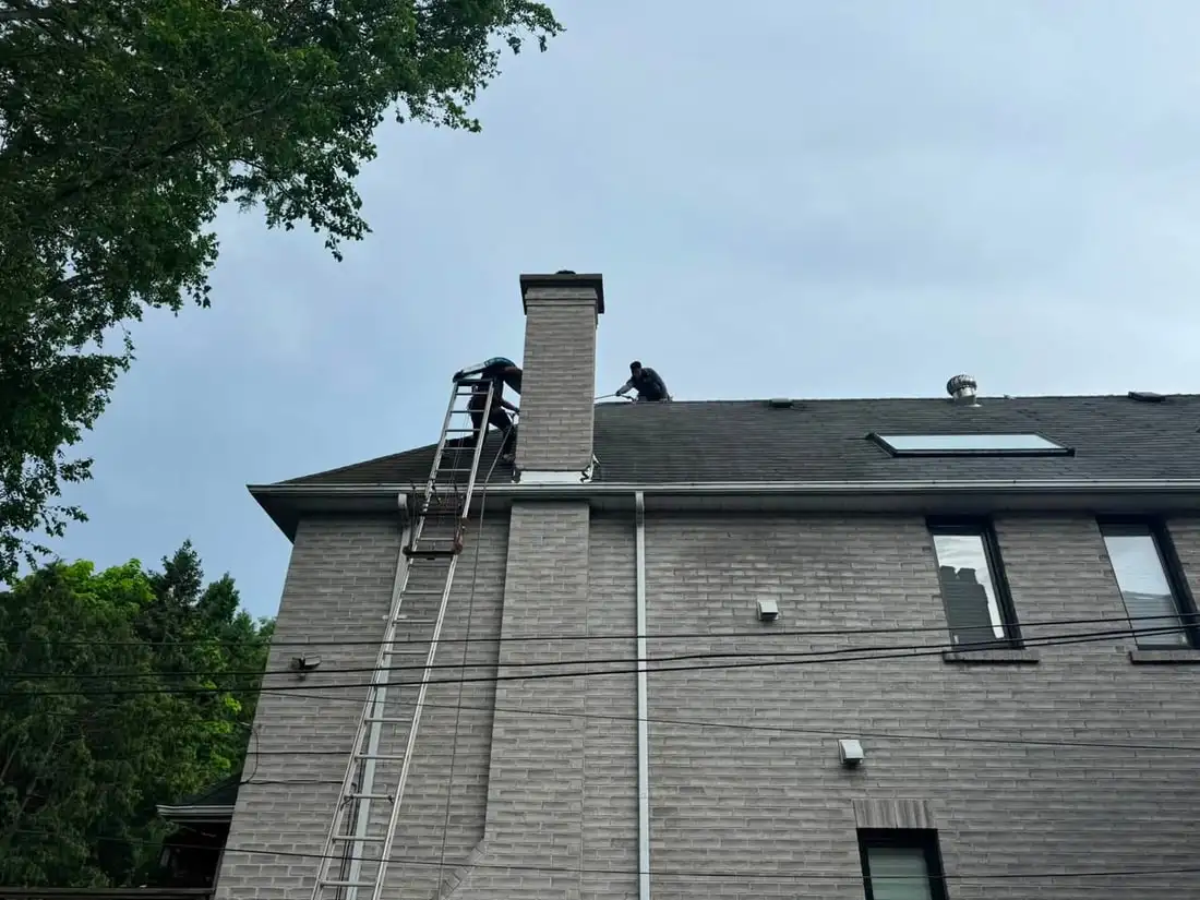 Looking up at a grey brick two-storey from the ground, a ladder with hoist rail running to the eave beside a tall chimney, two crew on the roof edge.