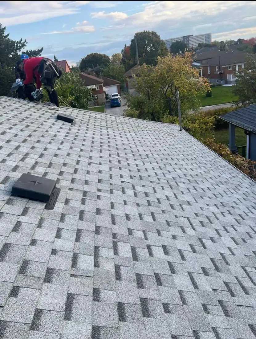 A light grey shingle roof with two crew members working at the ridge line, autumn trees and a residential street behind.