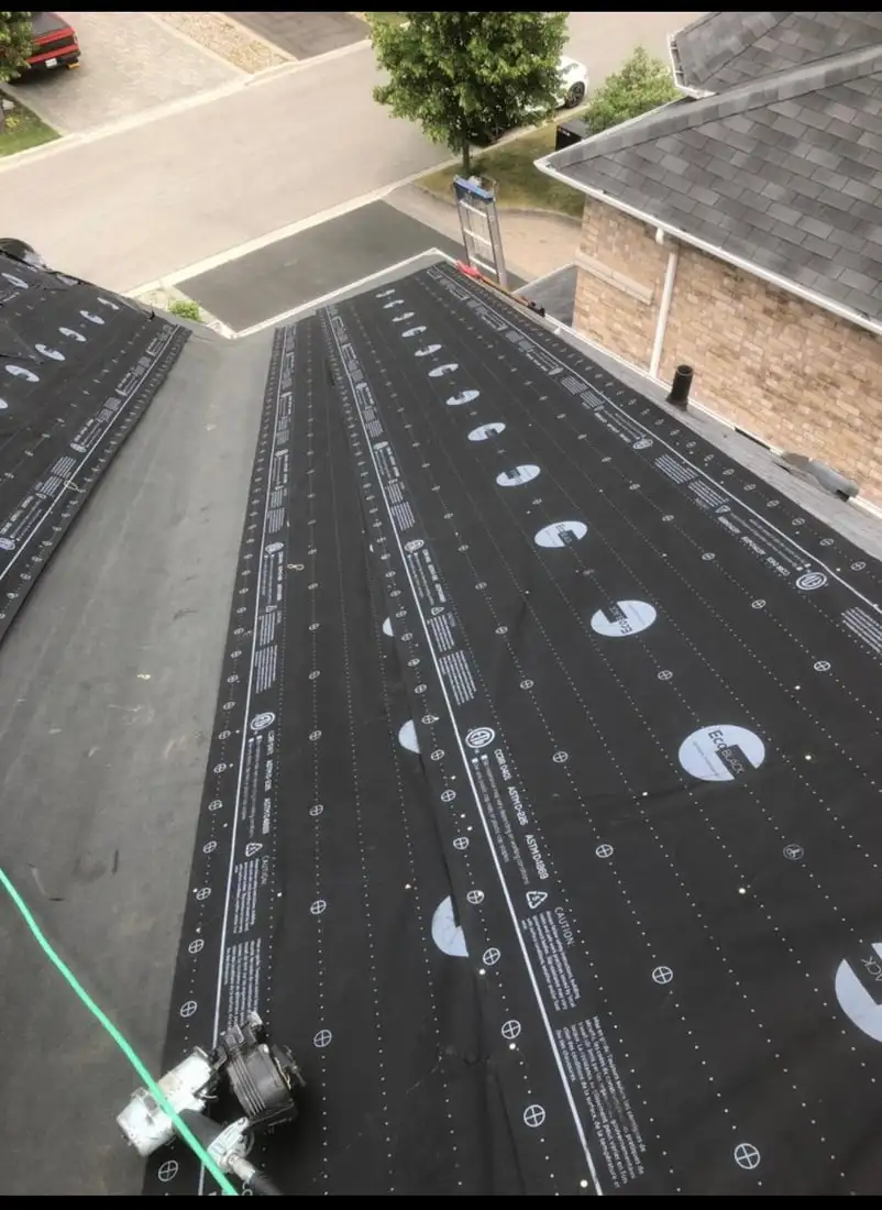 A roof deck stripped bare and fully covered in black synthetic underlayment with printed nailing lines, a coil nailer resting at the eave.