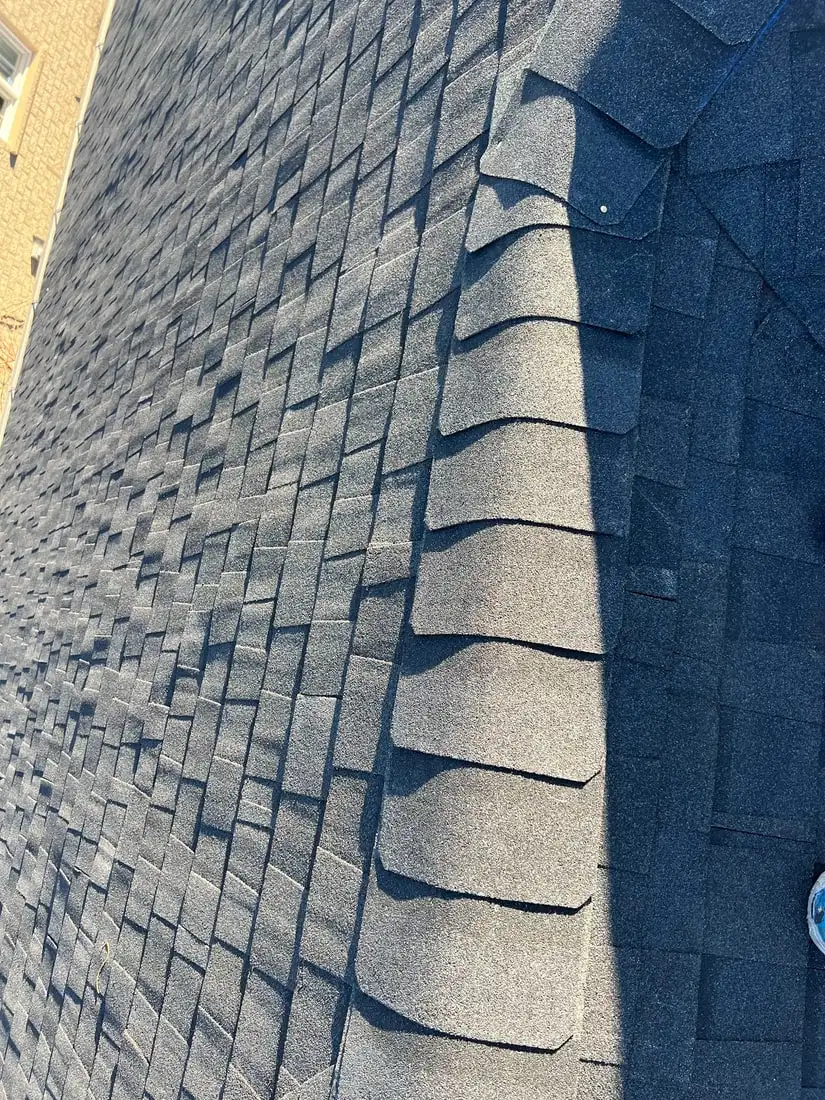 A tight detail of ridge cap shingles stepping down a hip line with deep shadow definition, brick wall at the edge.