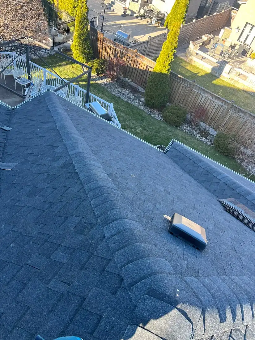 A freshly finished grey roof with new ridge cap, looking down past a white deck railing to a fenced backyard with cedars.