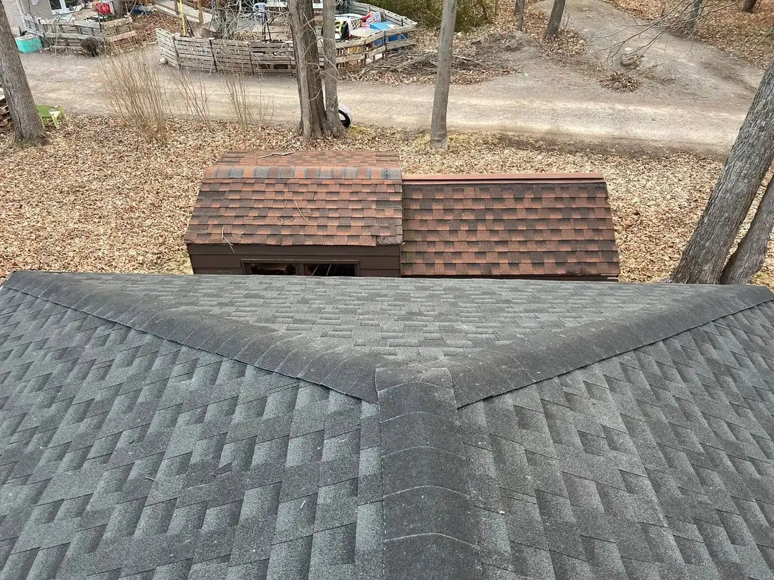 A freshly capped dark grey hip and ridge in the foreground, looking over a brown-shingled outbuilding to a wooded lot.