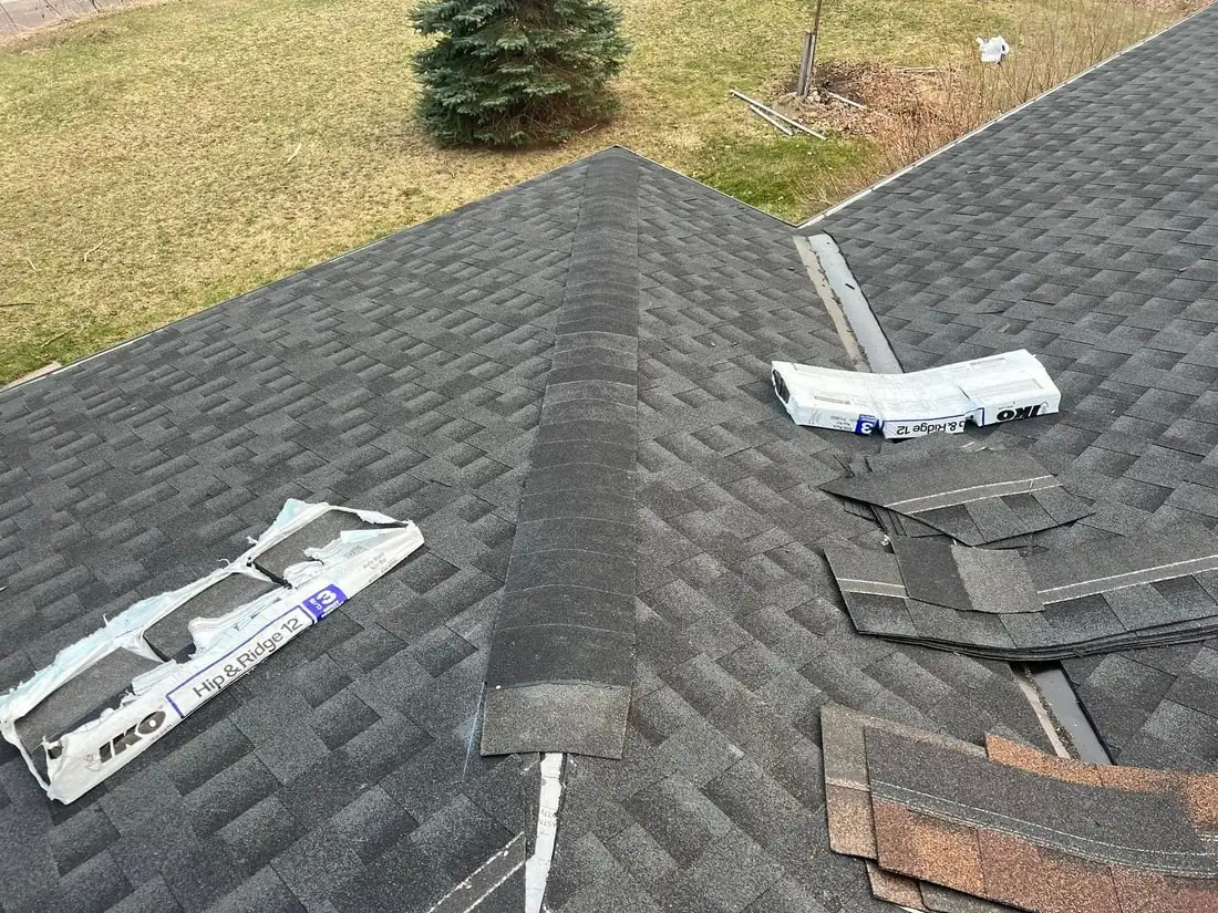 A new ridge cap running down the centre of a dark grey roof, empty IKO Hip and Ridge bundle wrappers and torn-off debris to one side.