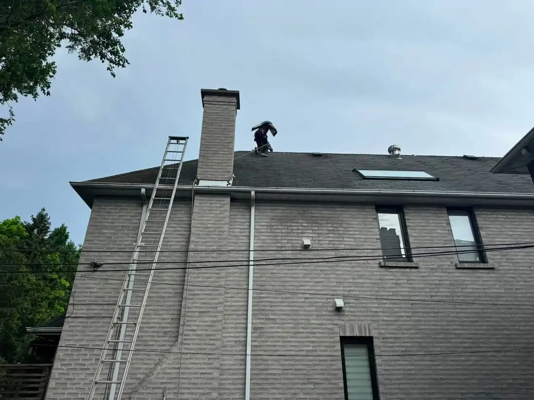A roofer in a red shirt carrying a shingle bundle on his shoulder along the ridge of a grey brick house.