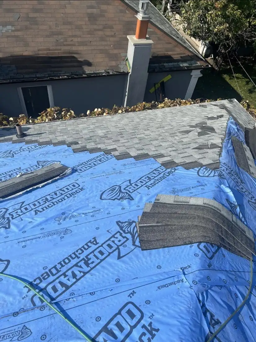A roof mid-job with blue Roofnado AnchorDeck underlayment laid over most of the deck and grey shingles partly installed at the top.