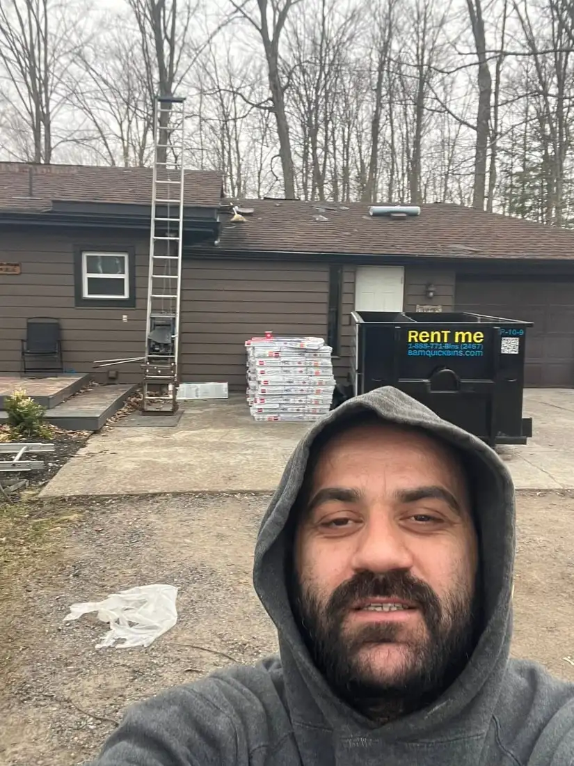 Viktor on a job site in a grey hoodie, with a ladder and shingle hoist up to the eave of a house and a rental bin behind him.