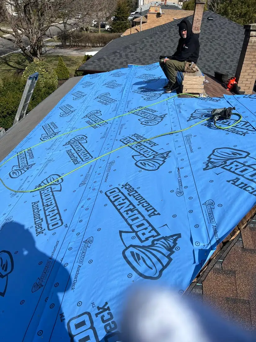 A worker sitting on the ridge above a full slope of blue Roofnado AnchorDeck underlayment with a chalk line snapped across it.